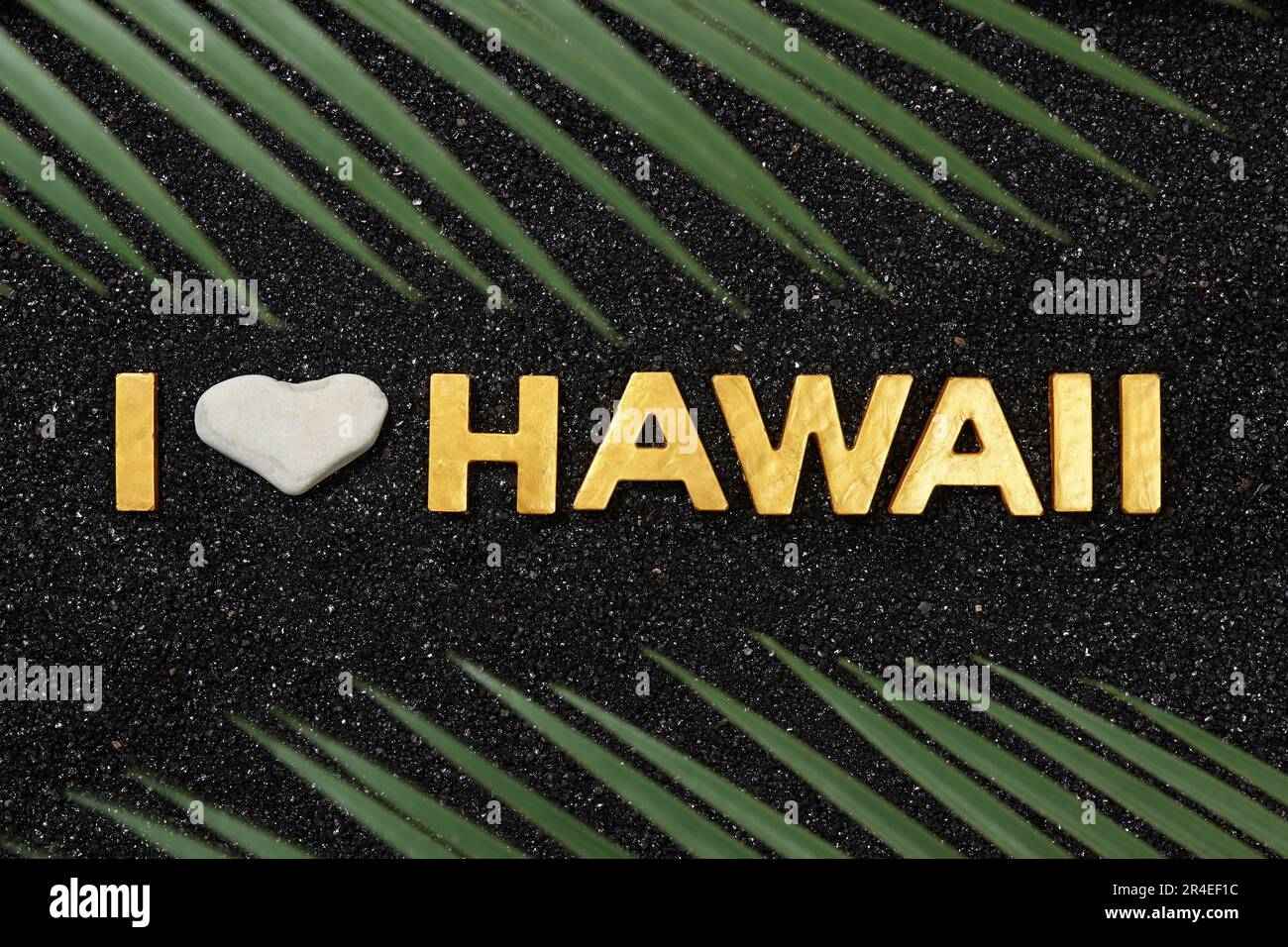 I Love Hawaii – Text im schwarzen Sandstrand und in Palmenblättern Stockfoto