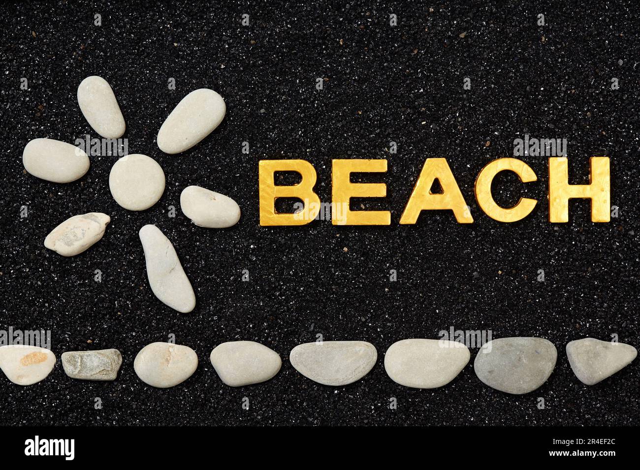 Strand - ein Wort in schwarzem Sand und ein Sonnensymbol mit Kieselsteinen Stockfoto