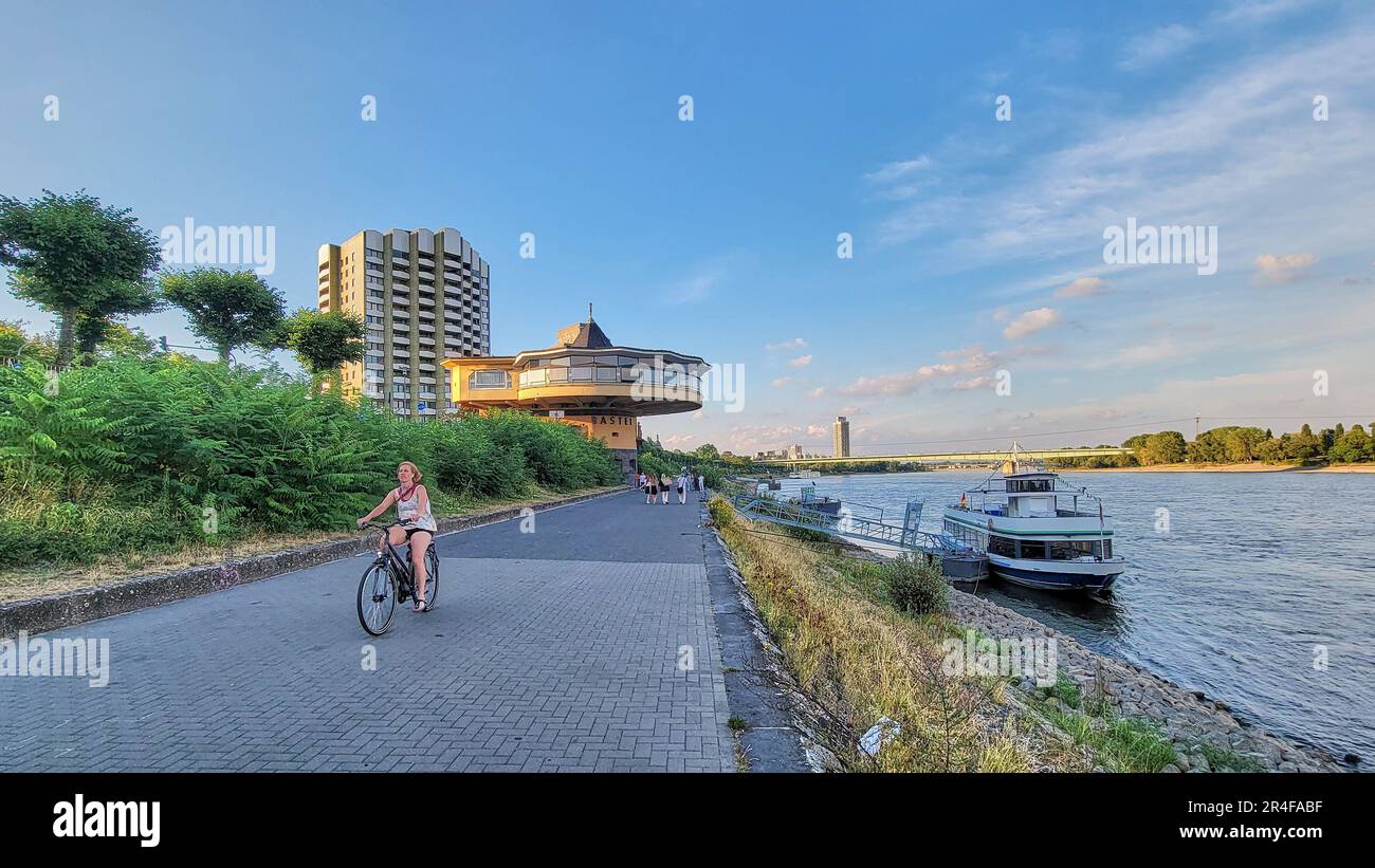 Frau radelt entlang der Rheinpromenade in Köln, mit dem berühmten Bastei-Architekturrestaurant im Art déco-Stil im Hintergrund. Stockfoto