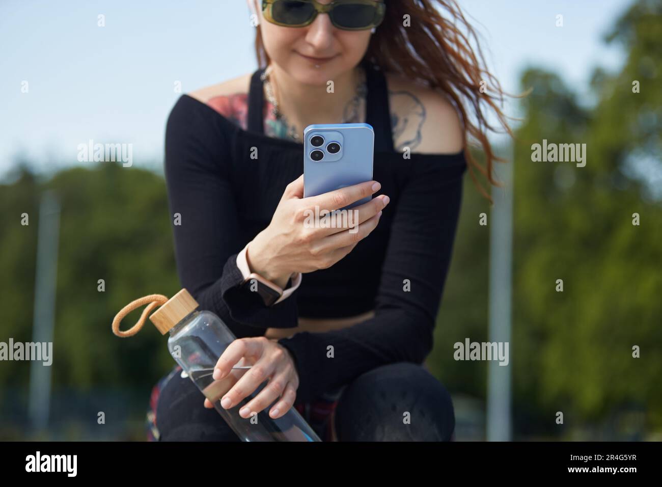 Skaterin sitzt in einem Skatepark mit einer wiederverwendbaren Glasflasche Wasser und Smartphone in den Händen. Vielfältige junge Frau mit tätowierter Brust mit einem M. Stockfoto