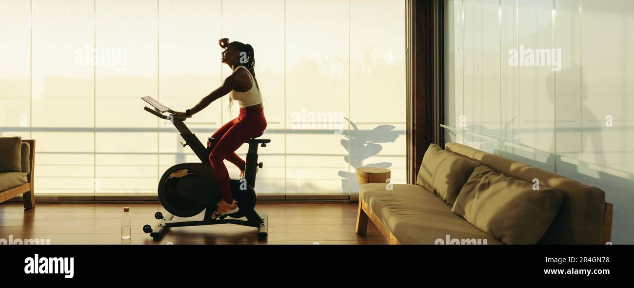 Afrikanische Frau verwendet zu Hause ein digitales Fitness-Fahrrad, um einen gesunden Lebensstil aufrechtzuerhalten. Sie wischte sich den Schweiß im Gesicht ab und raste mit dem Fahrrad weg Stockfoto