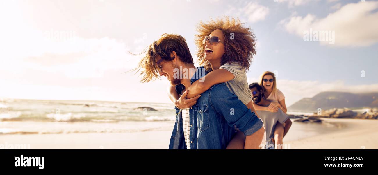 Glückliches Paar genießt einen sonnigen Tag am Strand, während der Mann seine Freundin mit dem Huckepack mitnimmt. Junge Liebhaber, die einen romantischen Sommerurlaub haben, m Stockfoto