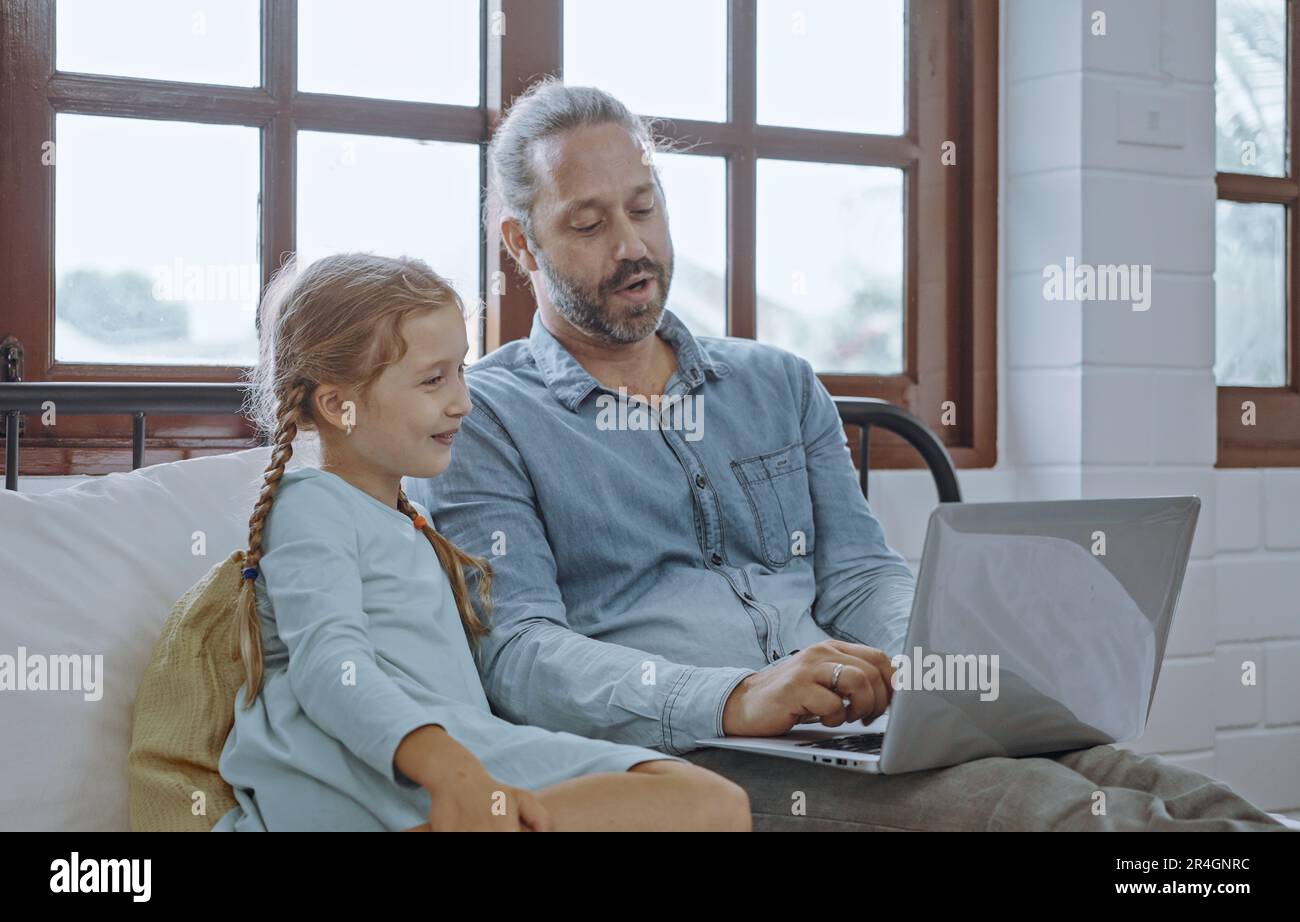 Vater benutzt Laptop-Computer mit der kleinen Tochter. Glückliche Familie, Vater entspannt sich und genießt es, zu Hause online zu arbeiten, während das kleine süße Mädchen im Bett liegt Stockfoto