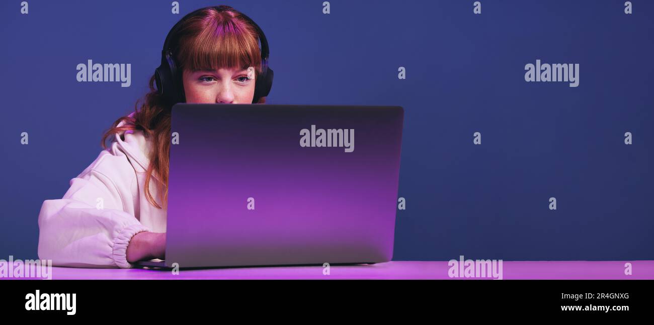 Junge Spielerin, die auf ihren Laptop-Bildschirm schaut, während sie ein Online-Videospiel in einem Studio spielt. Eine Frau mit Ingwerhaaren, die interaktives Streaming verwendet Stockfoto