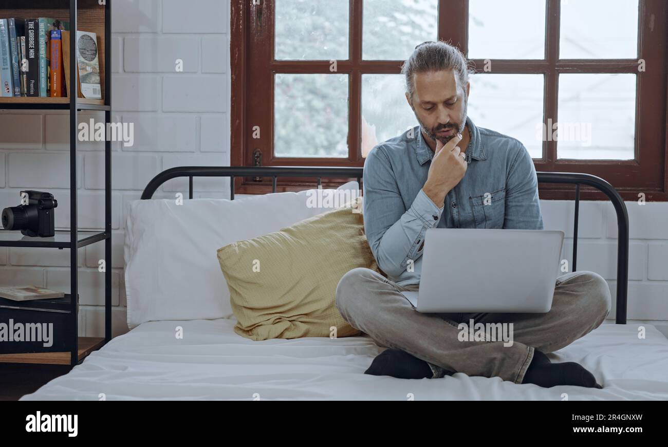 Ein junger besorgter Mann, der ein Notebook benutzt und im Schlafzimmer auf dem Bett sitzt. Stockfoto