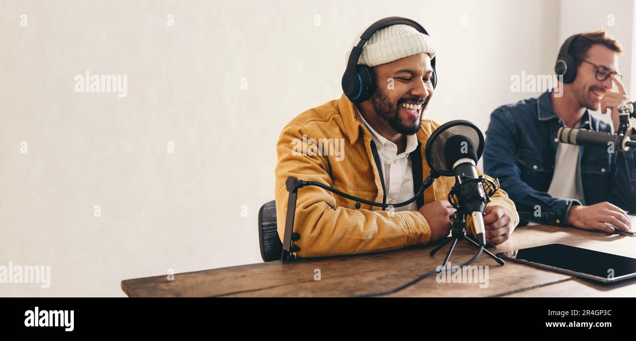 Ein Radio-dj lächelt, während er in einem Studio in ein Mikrofon spricht. Glücklicher junger Mann, der zusammen mit einem Gast eine Audioübertragung moderiert. Zwei junge Inhalte c Stockfoto