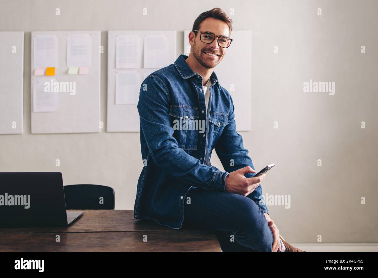Glücklicher junger Geschäftsmann, der in die Kamera lächelt, während er mit einem Smartphone auf dem Schreibtisch sitzt. Fröhlicher Geschäftsmann mit Brille, der aus der Ferne in einem cr arbeitet Stockfoto