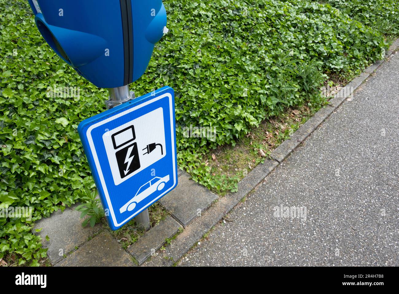 Blaues Schild mit einem Ladegerät darüber auf einem Parkplatz entlang eines grünen Gürtels, um anzuzeigen, dass es nur zum Aufladen von Elektroautos verwendet wird. Ladepunkt Elektrofahrzeuge Stockfoto