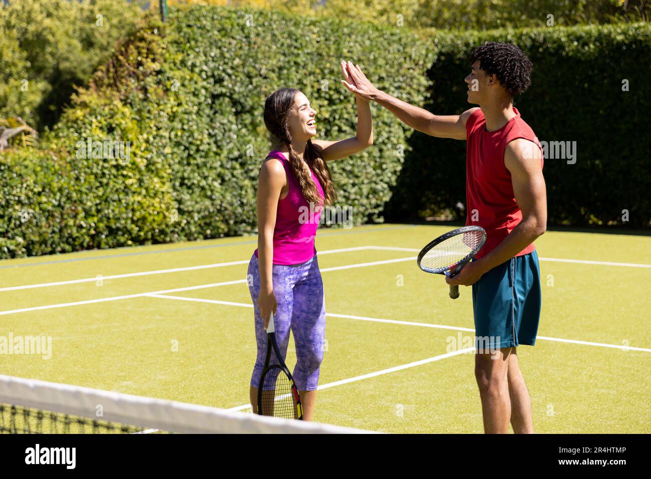 Ein fröhliches, birassisches junges Paar, das Tennisschläger hält und High Five gibt, während es im Gericht steht Stockfoto