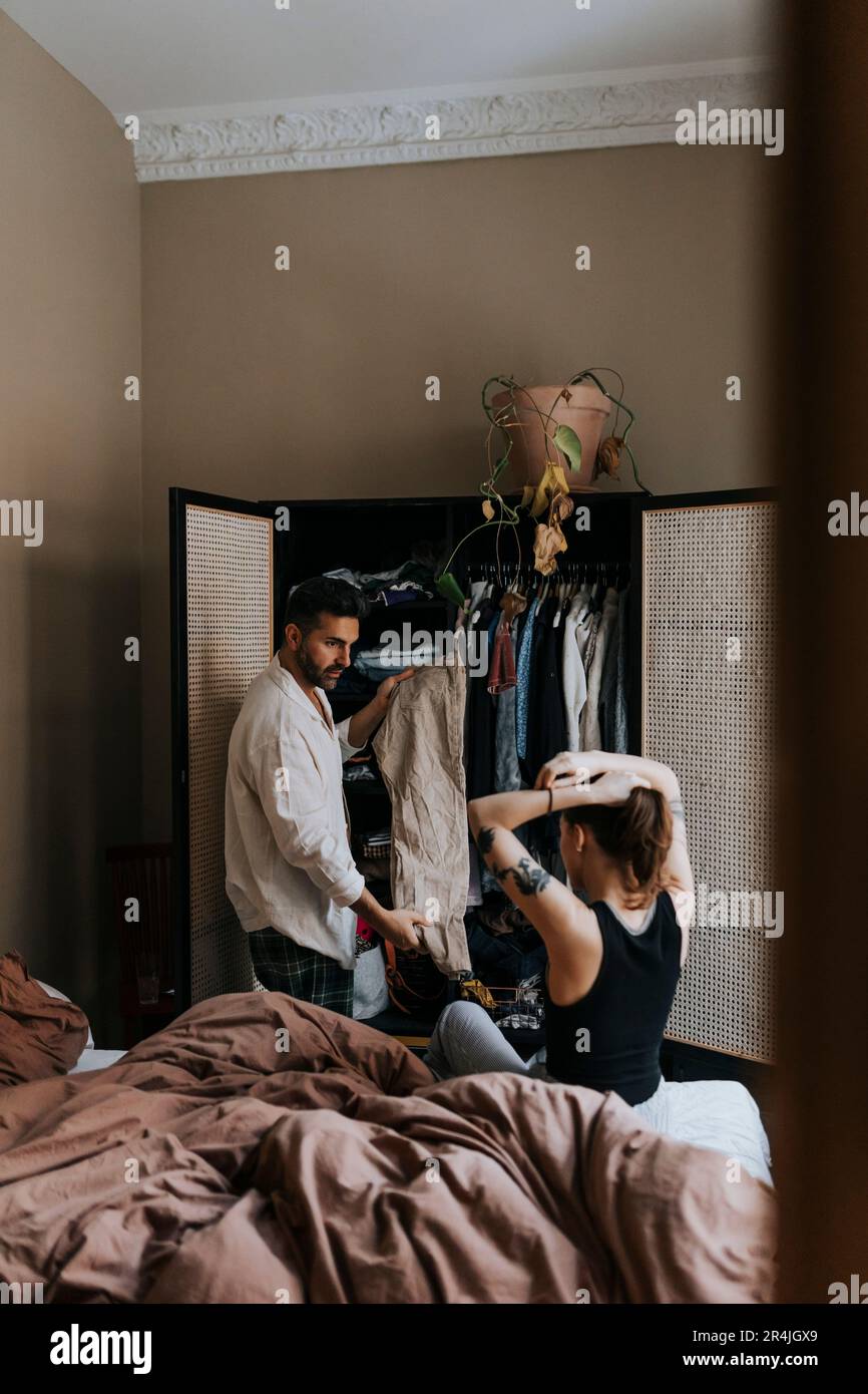 Ein Mann, der einer nicht binären Person Kleidung vorführt, während er zu Hause im Bett sitzt und Haare bindet Stockfoto