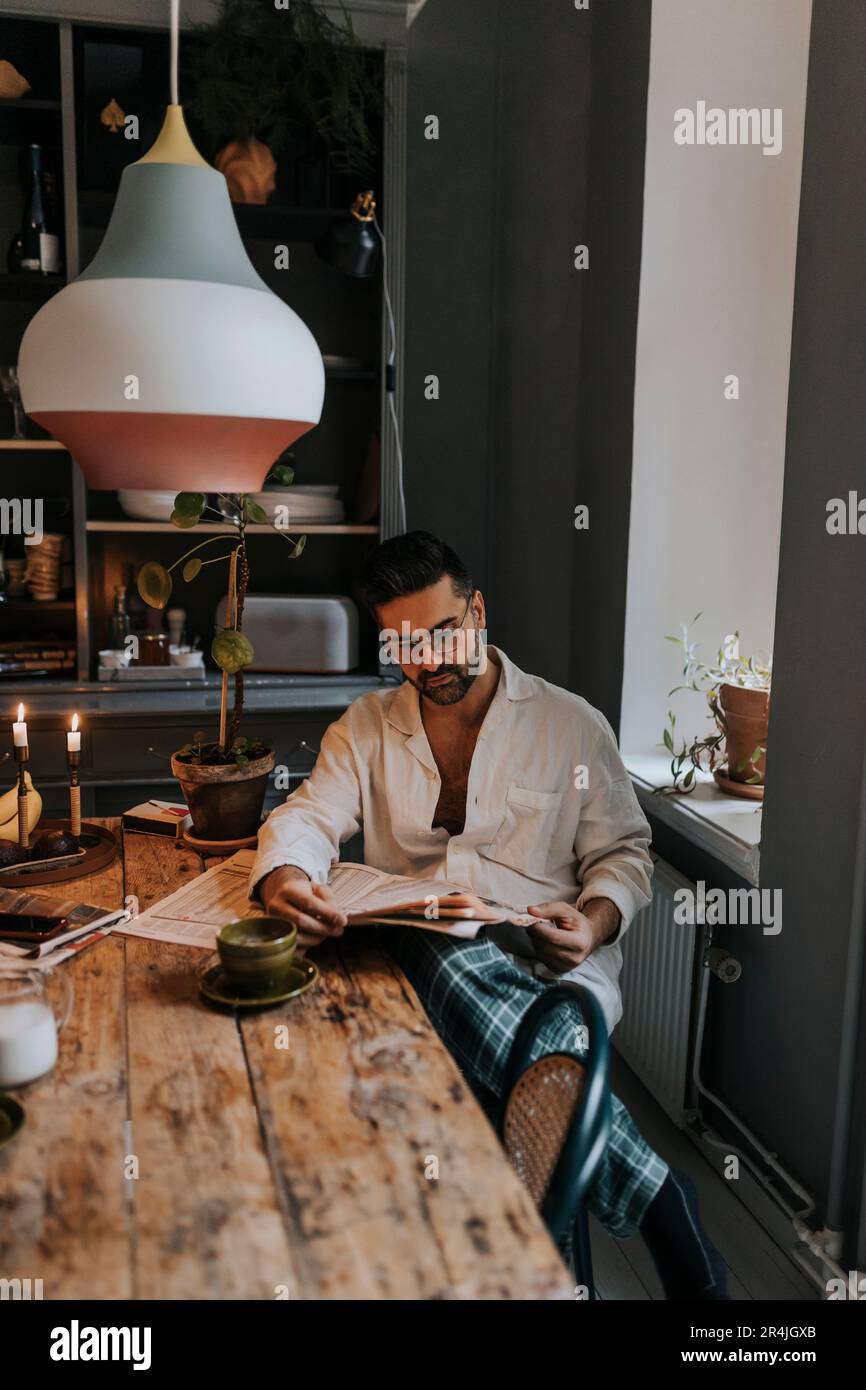 Mann liest Zeitung, während er zu Hause neben dem Tisch sitzt Stockfoto
