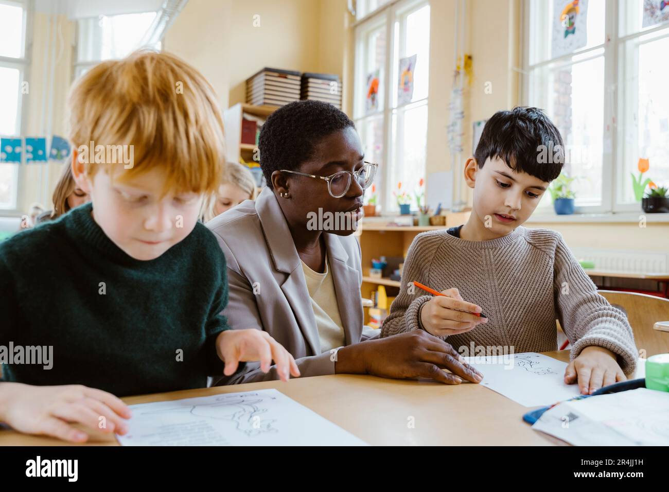 Lehrer, der Jungen beim Lösen von Problemen hilft, während sie im Klassenzimmer am Schreibtisch sitzen Stockfoto