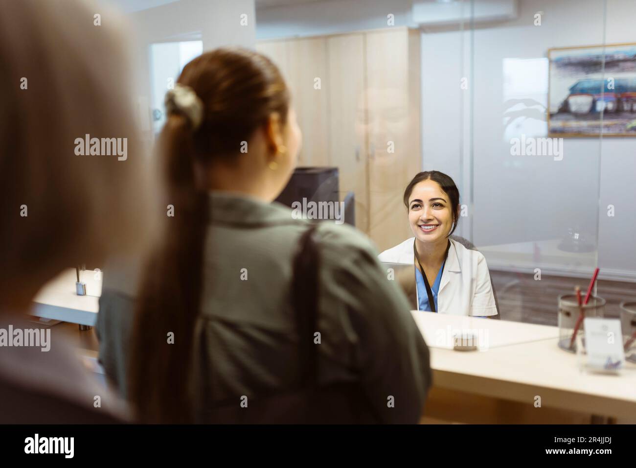 Lächelnde Rezeptionistin, die eine Frau durch einen Glasschirm im Krankenhaus ansieht Stockfoto