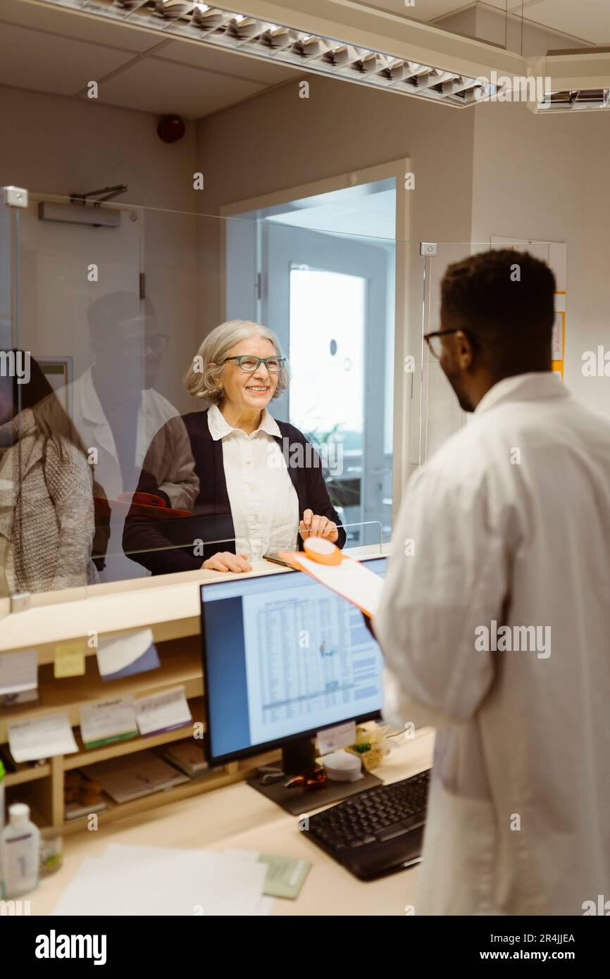 Lächelnde Seniorin, die mit dem männlichen Rezeptionisten über einen durchsichtigen Schild in der Klinik spricht Stockfoto