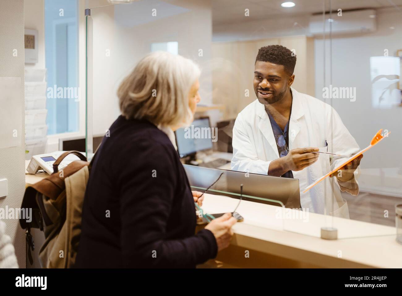 Lächelnder männlicher Rezeptionist hält Klemmbrett und spricht mit Seniorin durch durchsichtigen Schild in der Klinik Stockfoto