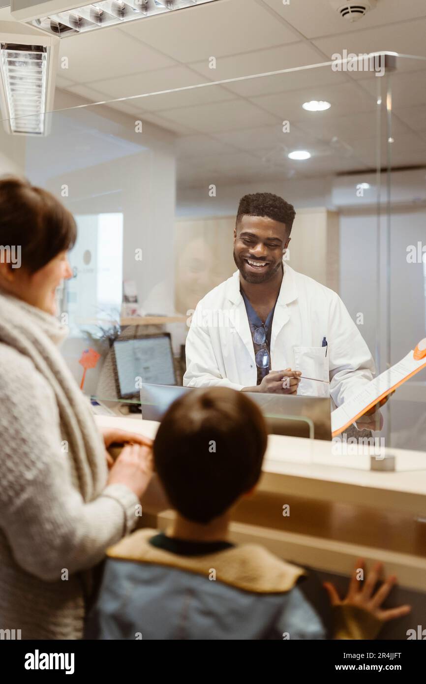 Lächelnder männlicher Empfangsmitarbeiter, der mit Mutter und Sohn über einen durchsichtigen Schild in der Klinik spricht Stockfoto