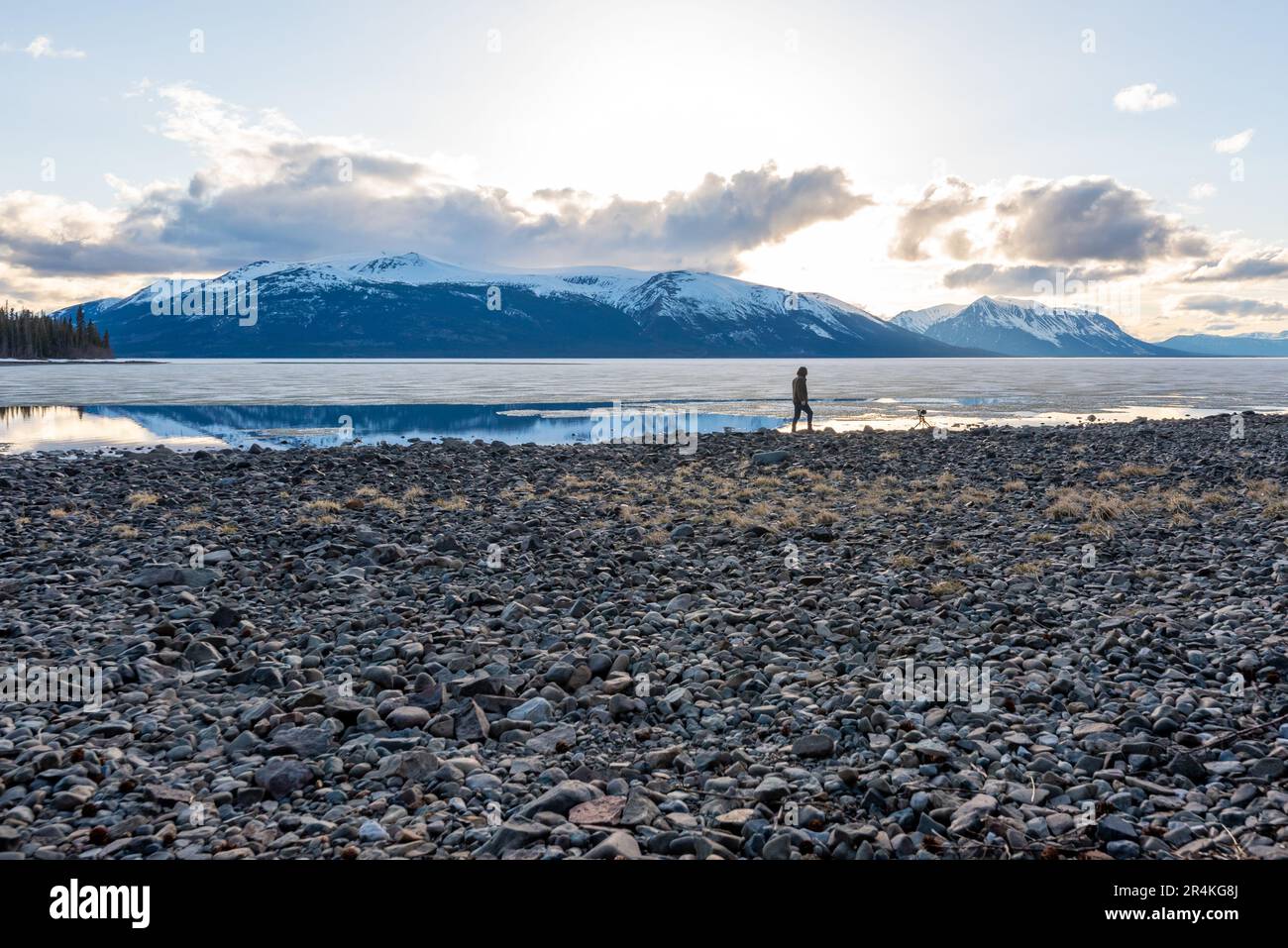 Ein Mann, der im Frühling neben einem teilweise gefrorenen, auftauenden See in Kanada steht, mit schneebedeckten Bergen im Hintergrund in der Wildnis. Stockfoto