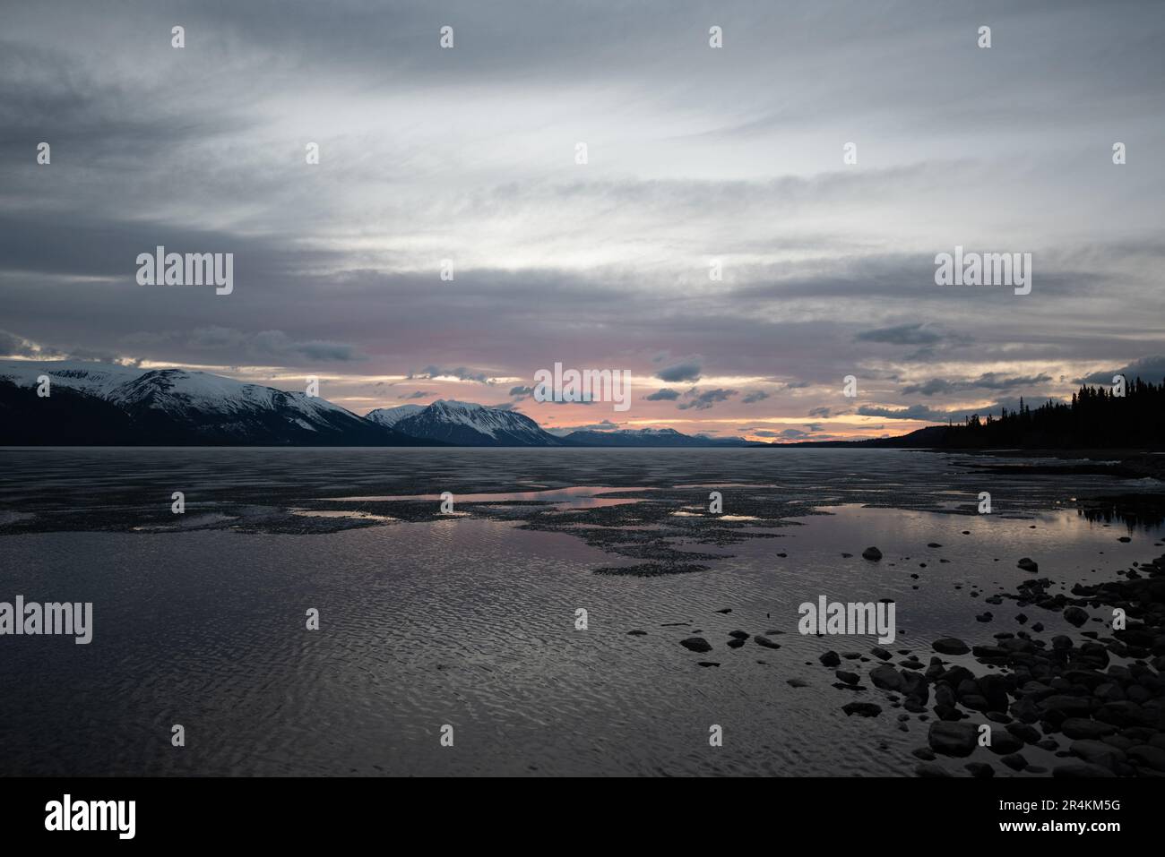 Sonnenuntergang am Rocky Lake in Atlin, British Columbia im Frühling. Schneebedeckte Berge spiegeln sich im ruhigen See mit dramatischem Himmel. Stockfoto