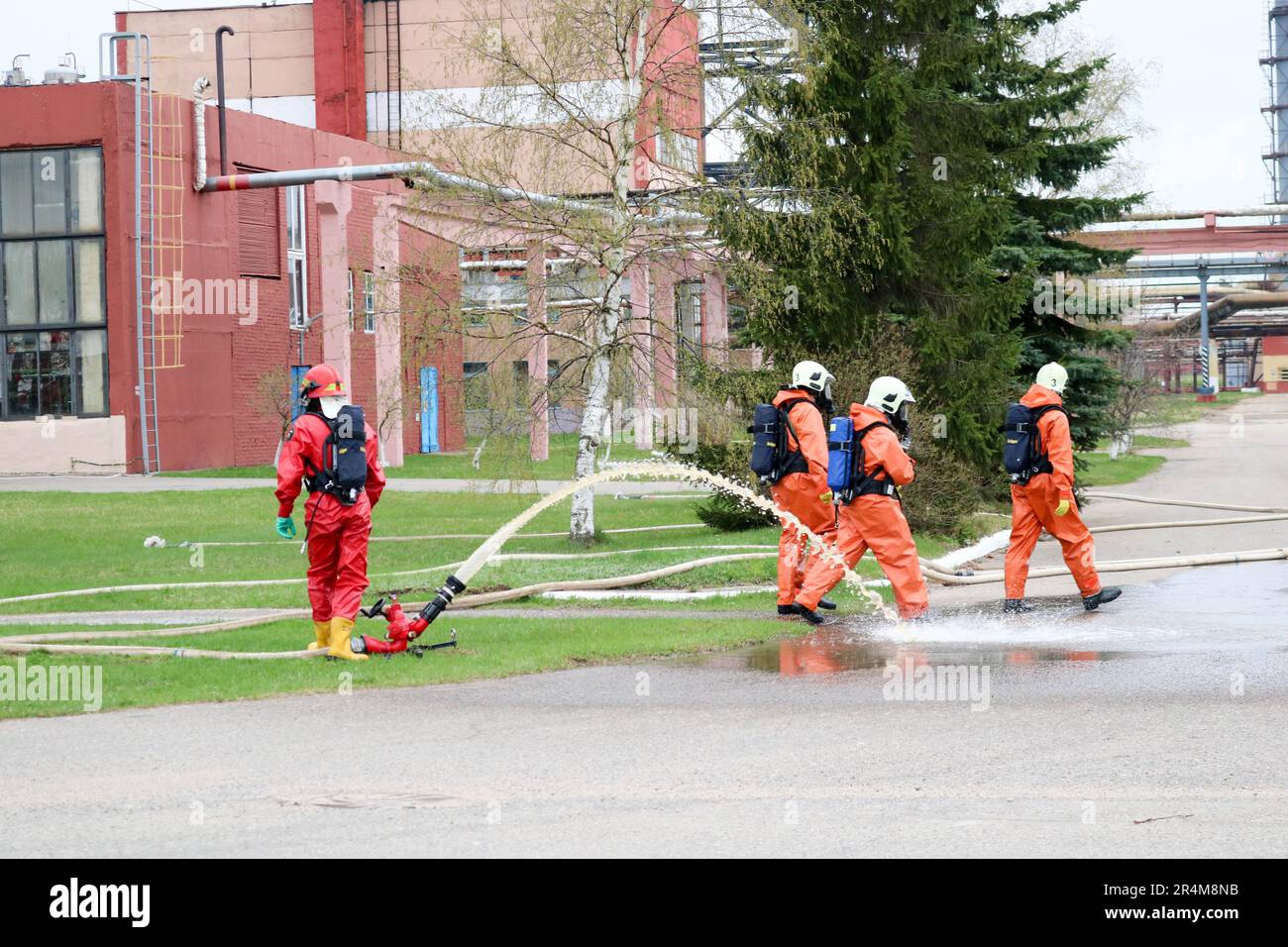 Professionelle Feuerwehrleute in orangefarbenen feuerfesten Anzügen in weißen Helmen mit Gasmasken testen Feuerlöschschläuche und Feuerwaffen, um einen Brand zu löschen Stockfoto