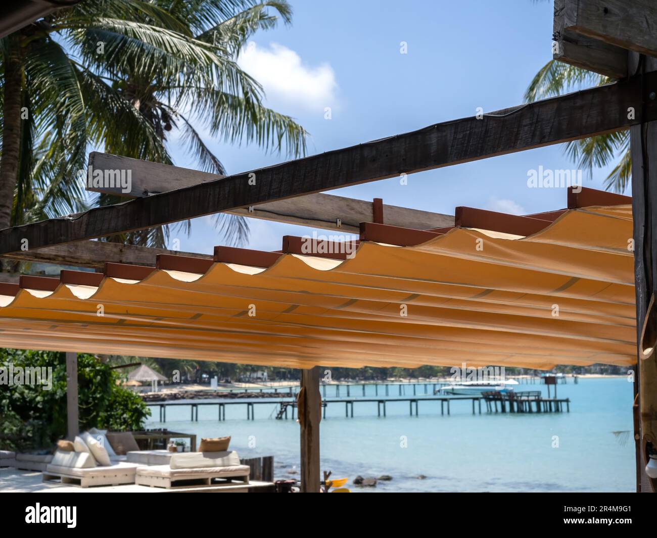 Ausziehbares Dach im Freien auf Holz- und Eisenkonstruktion am Strandrestaurant in der Nähe der tropischen Palmen und Meereslandschaft im Hintergrund. Foldabl Stockfoto