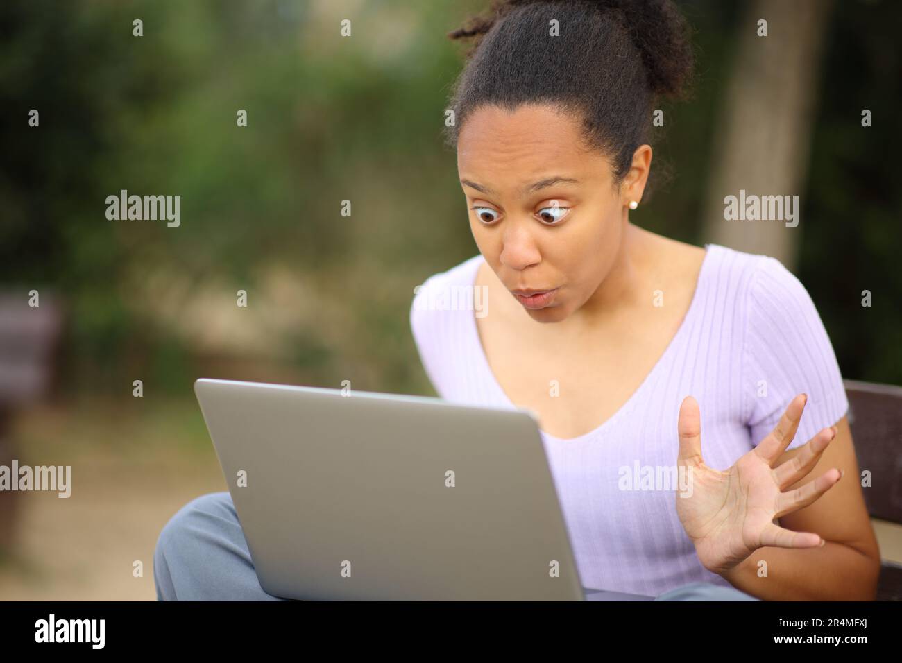 Überraschte schwarze Frau, die auf einer Bank im Park auf ihren Laptop-Inhalt schaut Stockfoto