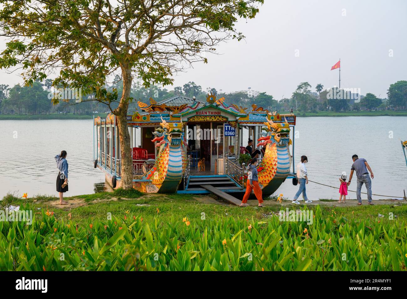 Ein Drachenboot, das auf dem Parfüm-Fluss vor Anker liegt, und eine einheimische Familie, die am Flussufer in Hue, Vietnam, den Alltag durchlebt. Stockfoto