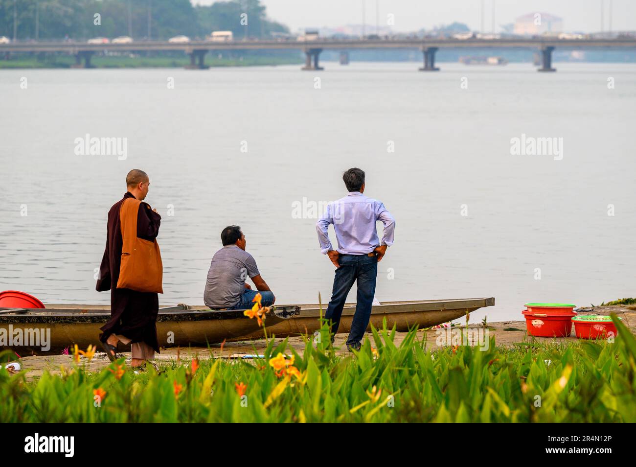 Ein buddhistischer Mönch wandelt von zwei Männern in stiller Unterhaltung und blickt auf den Perfume River im frühen Abendlicht, Hue, Vietnam. Stockfoto