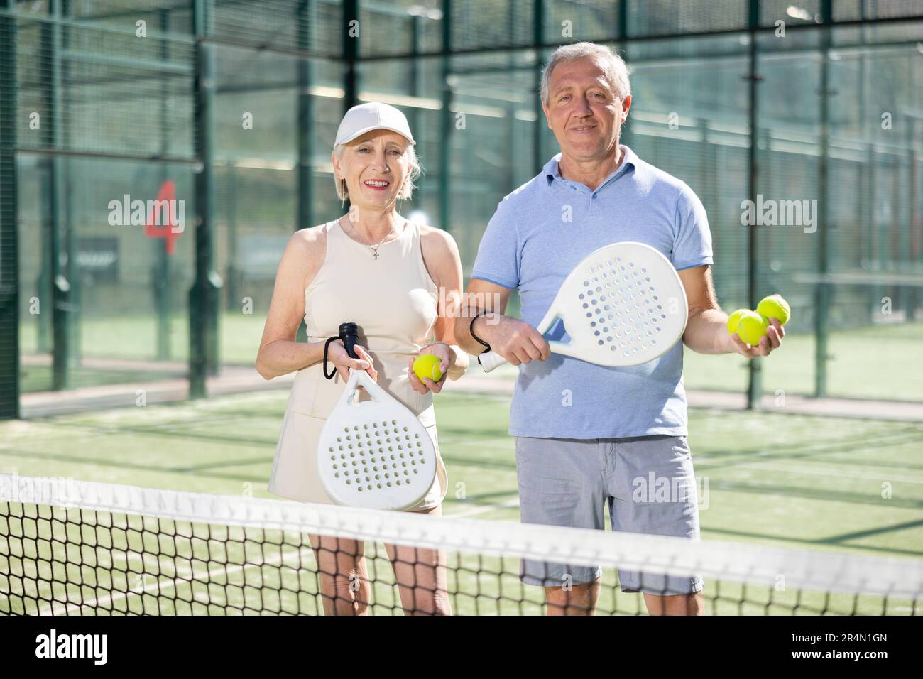 Alter Mann und Frau posieren mit Padel Schläger in den Händen neben dem Netz auf dem Tennisplatz Stockfoto
