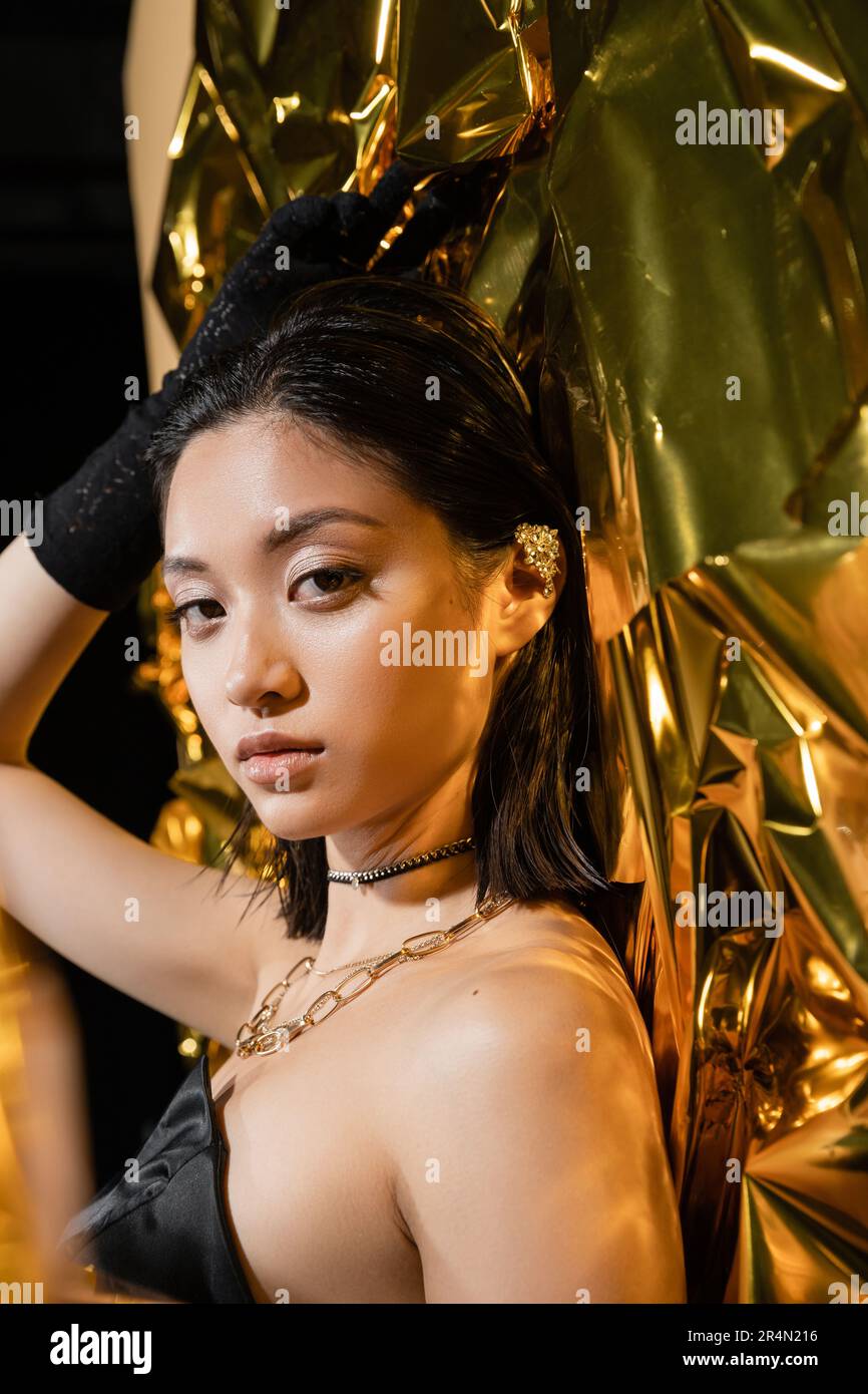 Elegante junge asiatische Frau mit nasser Frisur und kurzen Haaren in trägerlosem Kleid mit schwarzem Handschuh, während sie neben goldenem Hintergrund steht, Model Stockfoto
