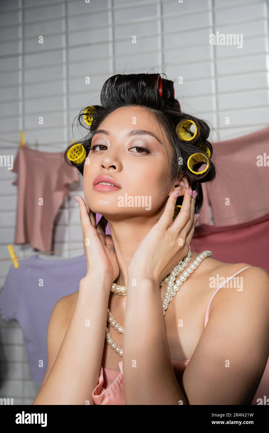 Junge asiatische Frau, die Haare mit Lockenwicklern berührt und in Perlenkette posiert, in der Nähe von sauberer und nasser Wäsche, die auf verschwommenem Hintergrund hängt, Hausarbeit, Hure Stockfoto