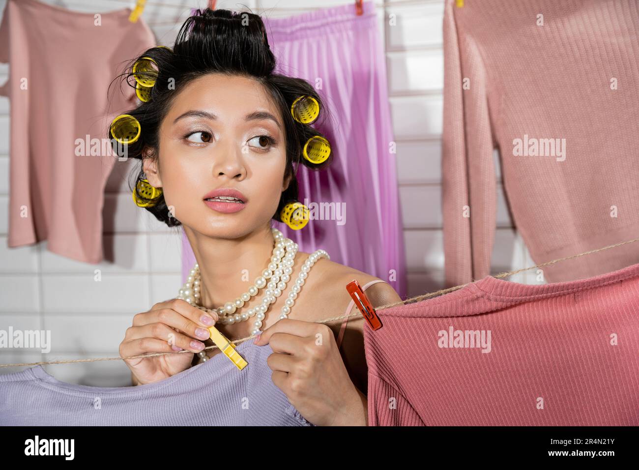 Nachdenkliche junge asiatische Frau mit Lockenstäben und Perlenkette, die Wäscheklammer hält und zu Hause saubere und nasse Wäsche mit verschwommenem Hintergrund aufhängt, Stockfoto