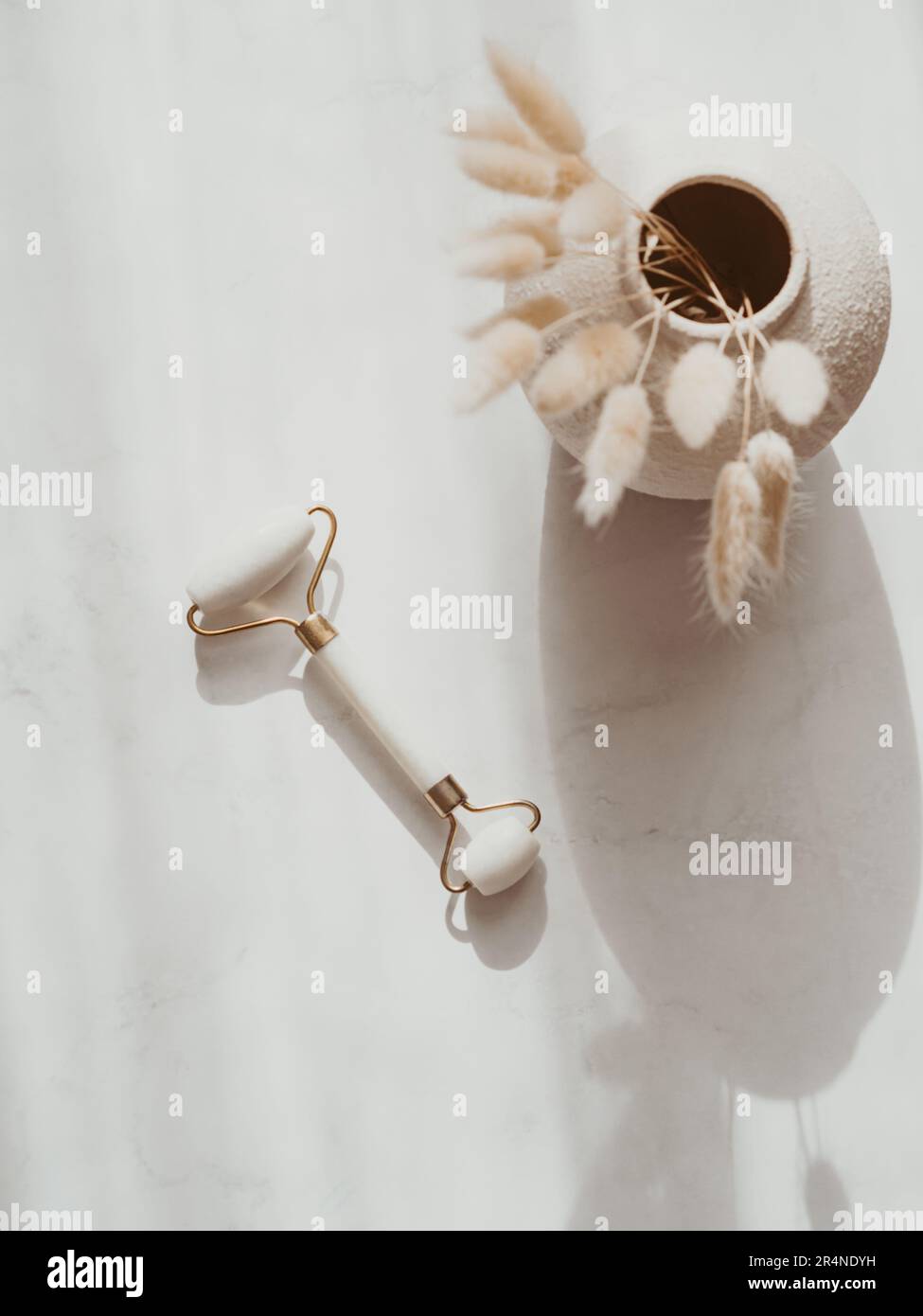 Weißer Jade-Gesichtsroller für Schönheitsmassagen und trockene Blumen in Vase auf weißem Hintergrund mit Schatten. Draufsicht. Speicherplatz kopieren. Stockfoto