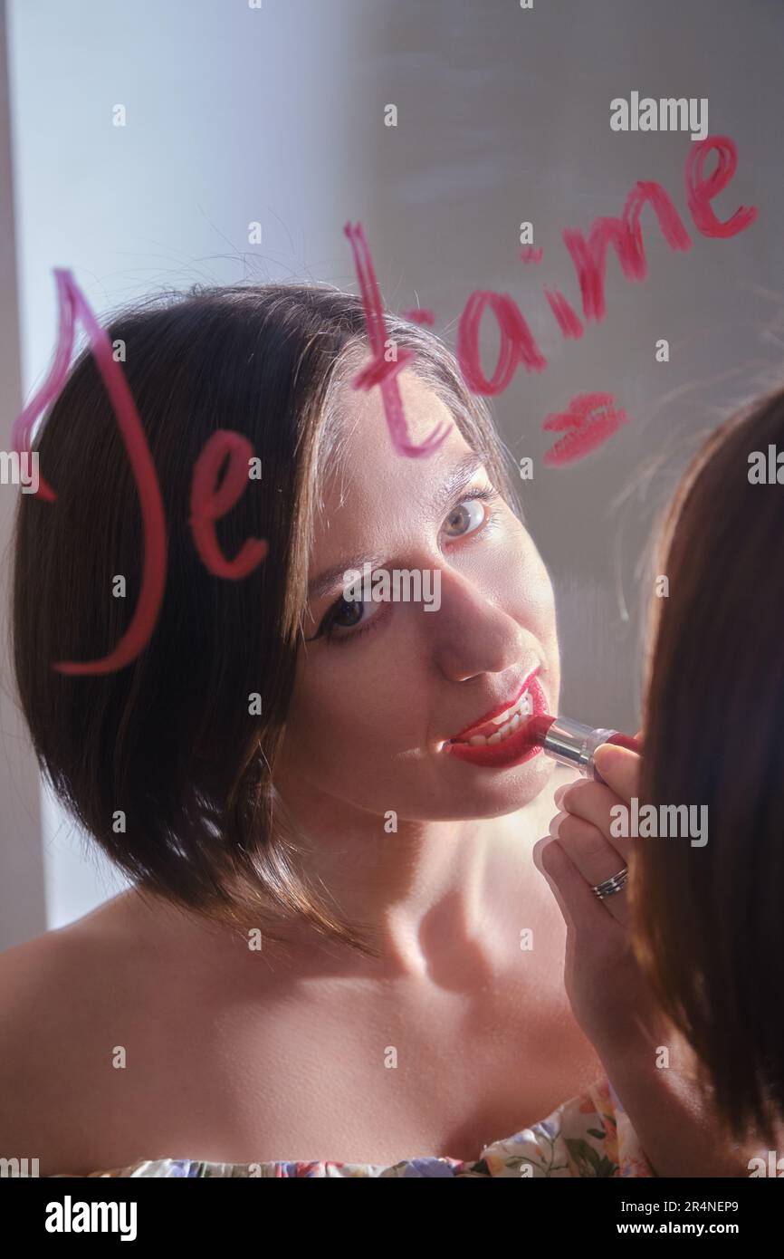 Eine glückliche Weiße sieht durch ihr Spiegelbild in die Kamera und trägt ihren Lippenstift auf. Auf dem Spiegel steht "je Taime", eine Nahaufnahme Stockfoto