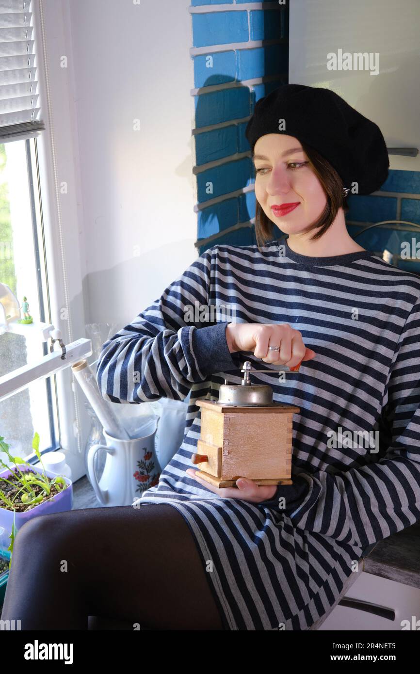 Glückliche junge Frau mahlt Kaffee mit einer handgemachten alten Mühle in der Küche und schaut in die Kamera Stockfoto