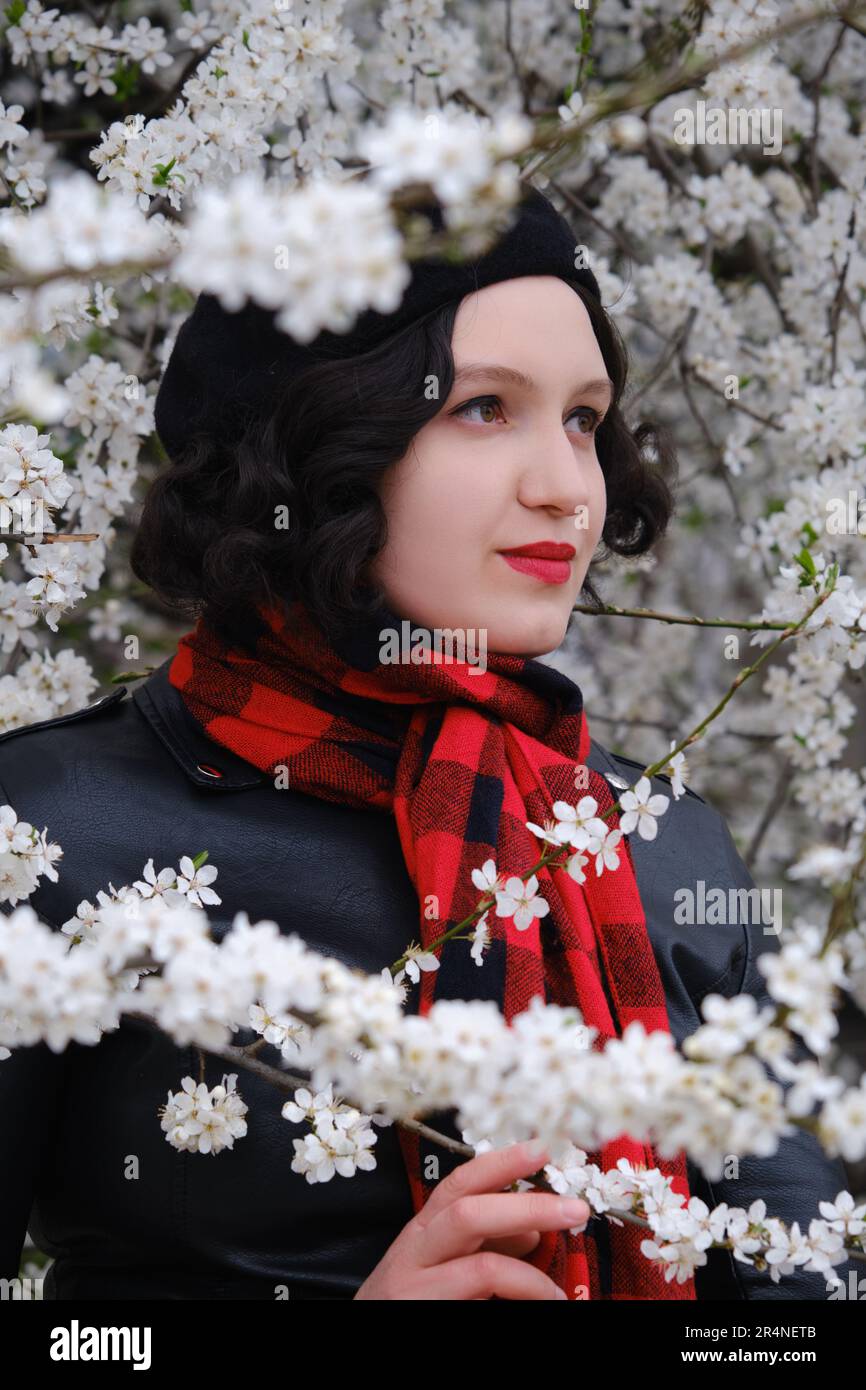 Wunderschöne junge Frau in Ledermantel und rotem Schal nahe der Kirschblüte, Nahaufnahme des Porträts Stockfoto