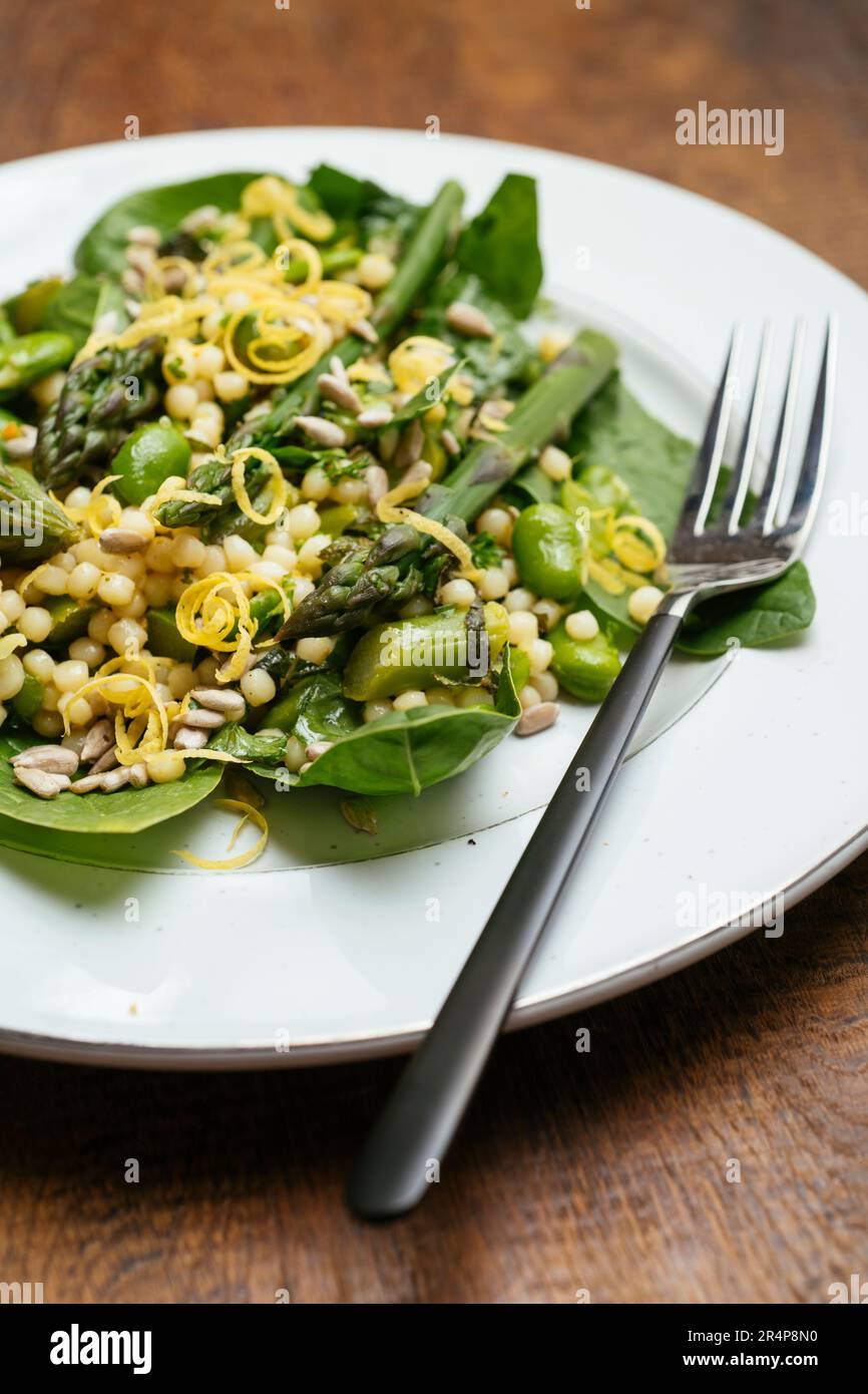 Teller mit einem hausgemachten veganen Couscous-Salat im Frühling mit grünem Spargel, Favabohnen und Baby-Spinat. Stockfoto