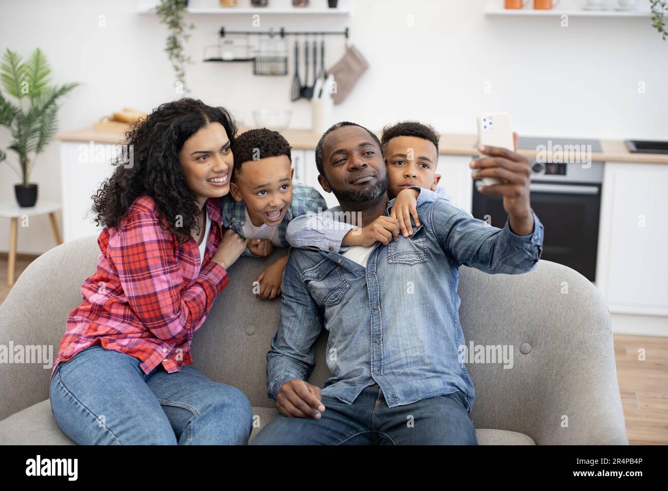 Positive multikulturelle vierköpfige Familie, die ein modernes Smartphone benutzt, während sie sich im Wohnzimmer ausruht. Liebenswerte Söhne, die Eltern umarmen, während Dad Selfie über mobile Webcam im Zimmer macht. Stockfoto
