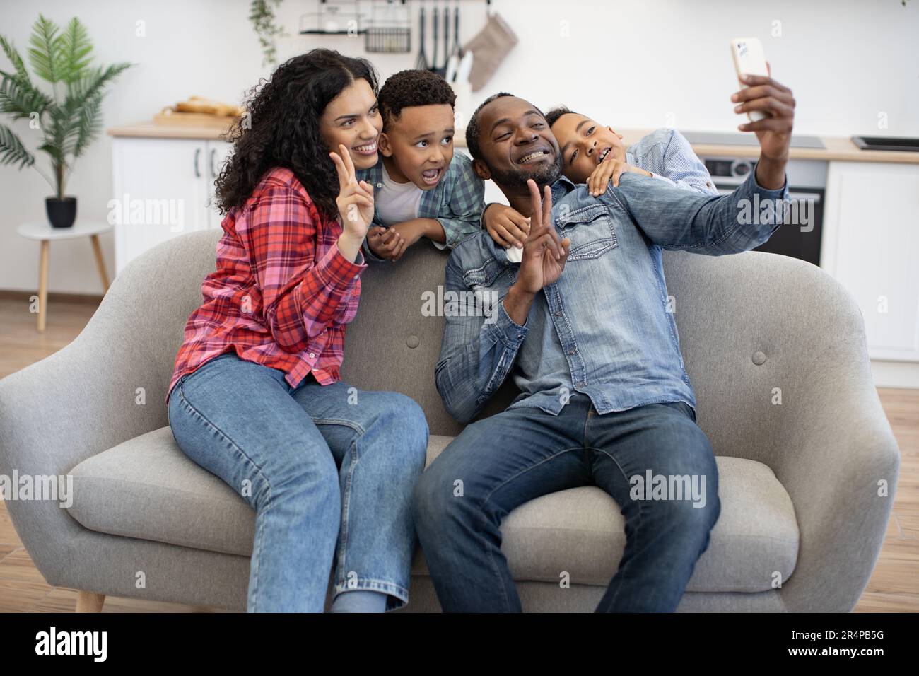 Ein multikultureller Vater mit Telefon und Mutter mit V-Schild, während energiegeladene Kinder hinter dem Sofa in der Lounge des Zuhauses stehen. Glückliches Paar mit Söhnen, das sich selbst über die mobile App fotografieren lässt. Stockfoto