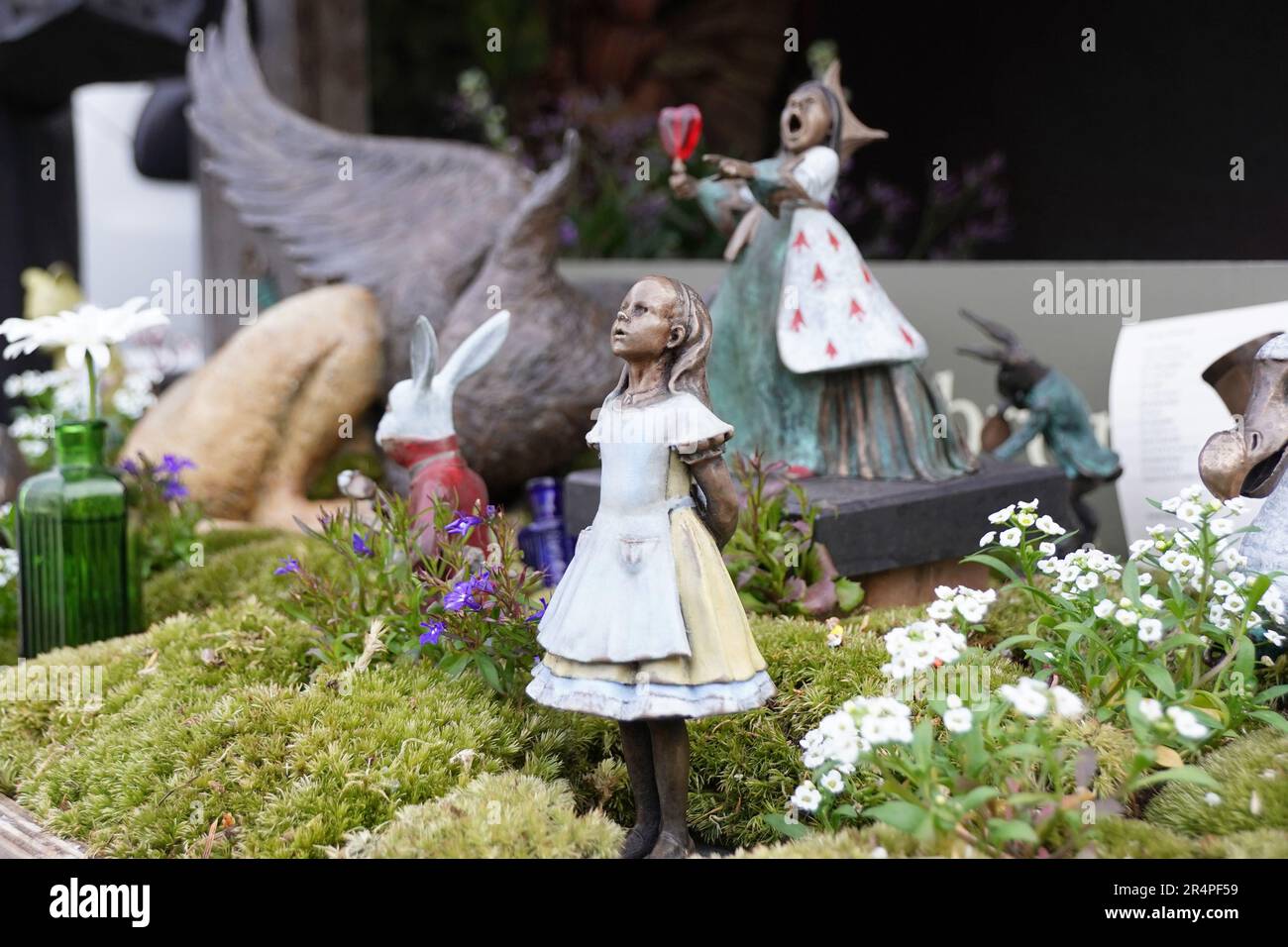 Eine Landszene von Alice in Wonder bei der Chelsea Flower Show, Alice mit Blick nach oben und die Herzkönigin im Hintergrund mit einem Glas Wein. Stockfoto