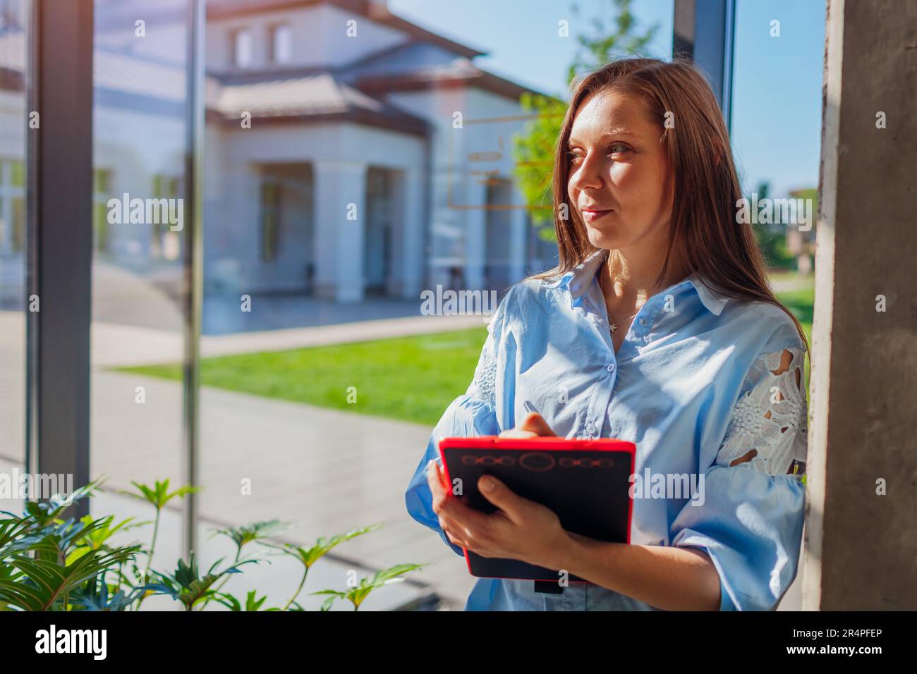Zufriedener, sicherer Büroangestellter, der das Tablet am Fenster hält. Eine weiße junge Frau, die in die Kamera schaut. Geschäftskonzept. Leerzeichen Stockfoto