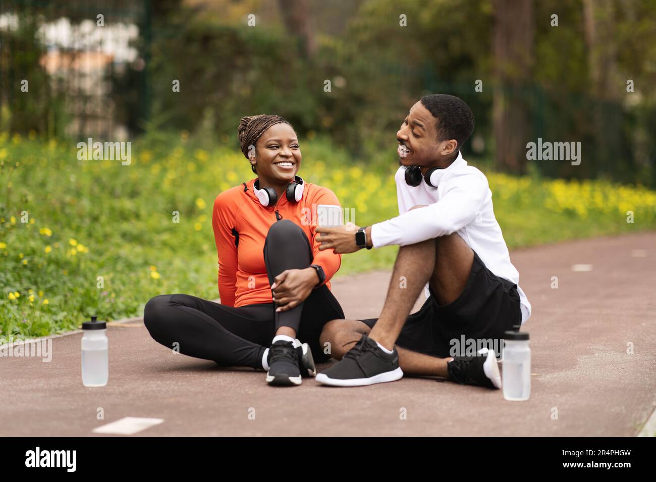 Glückliches, junges schwarzes Paar sitzt auf der Straße mit Wasserflaschen, ein Mann empfahl einen Sport-Blog am Telefon Stockfoto