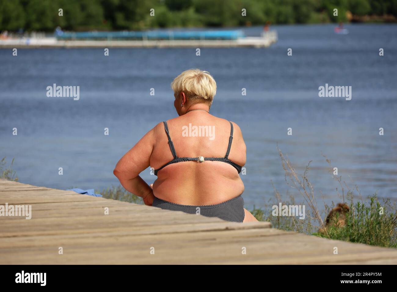 Übergewichtige Frau im Badeanzug, die auf dem Wasserhintergrund sitzt. Sommerurlaub am Fluss Strand, Überhitzung und Gewichtsverlust Konzept Stockfoto