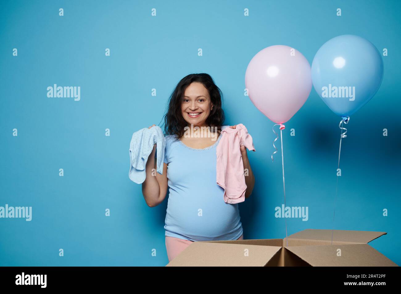 Die schöne, braune Frau lächelt, schaut in die Kamera, hält die Bodys von Neugeborenen auf einem isolierten blauen Hintergrund Stockfoto