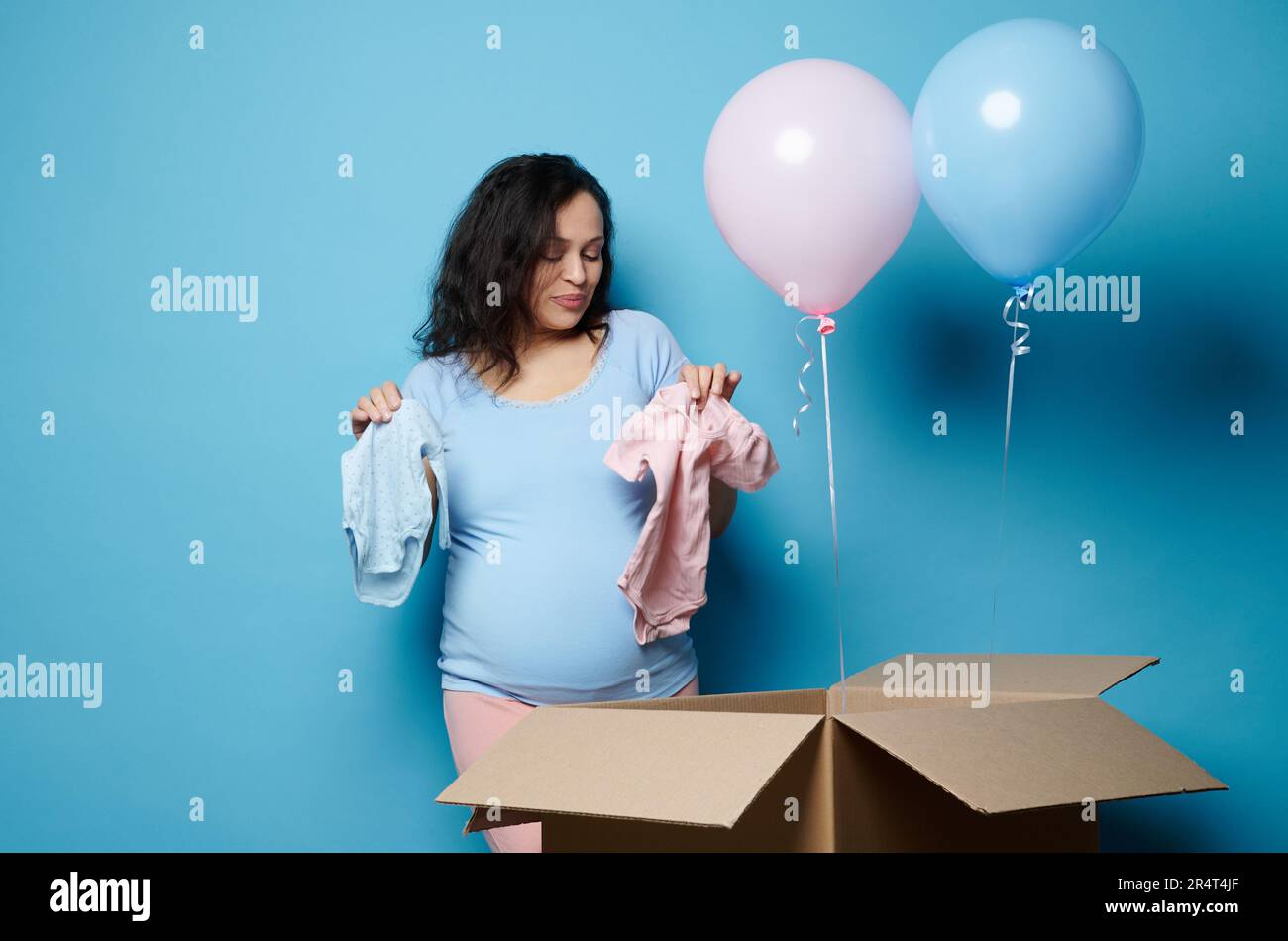 Schwangere Frau, schwangere Mutter, die blaue und pinkfarbene Neugeborene-Bodys aus einer Pappschachtel auf einer Gender-Enthüllungs-Party herausnimmt Stockfoto