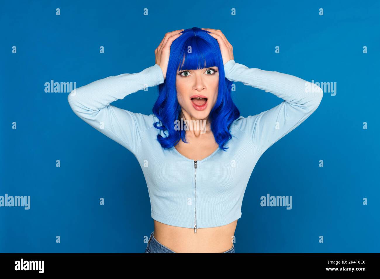 Schockiertes junges weibliches Model mit gefärbtem, hellblauem Haar in legerer Kleidung, das den Kopf packt und mit offenem Mund in die Kamera schaut Stockfoto