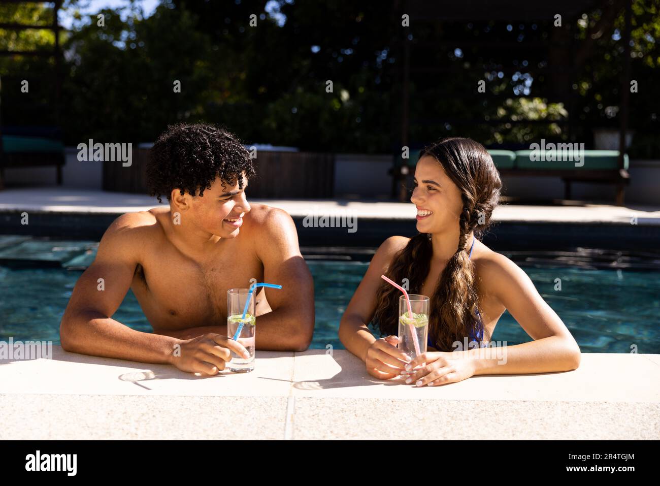 Ein birassisches junges Paar mit Getränken, das sich gegenseitig anschaut, während es am sonnigen Tag im Swimmingpool genoss Stockfoto