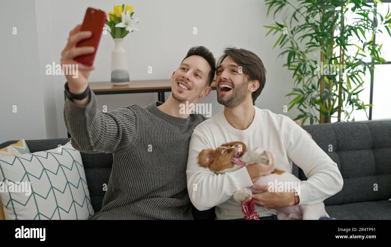 Zwei Männer machen Selfie mit dem Smartphone und sitzen zu Hause auf dem Sofa mit dem Hund Stockfoto