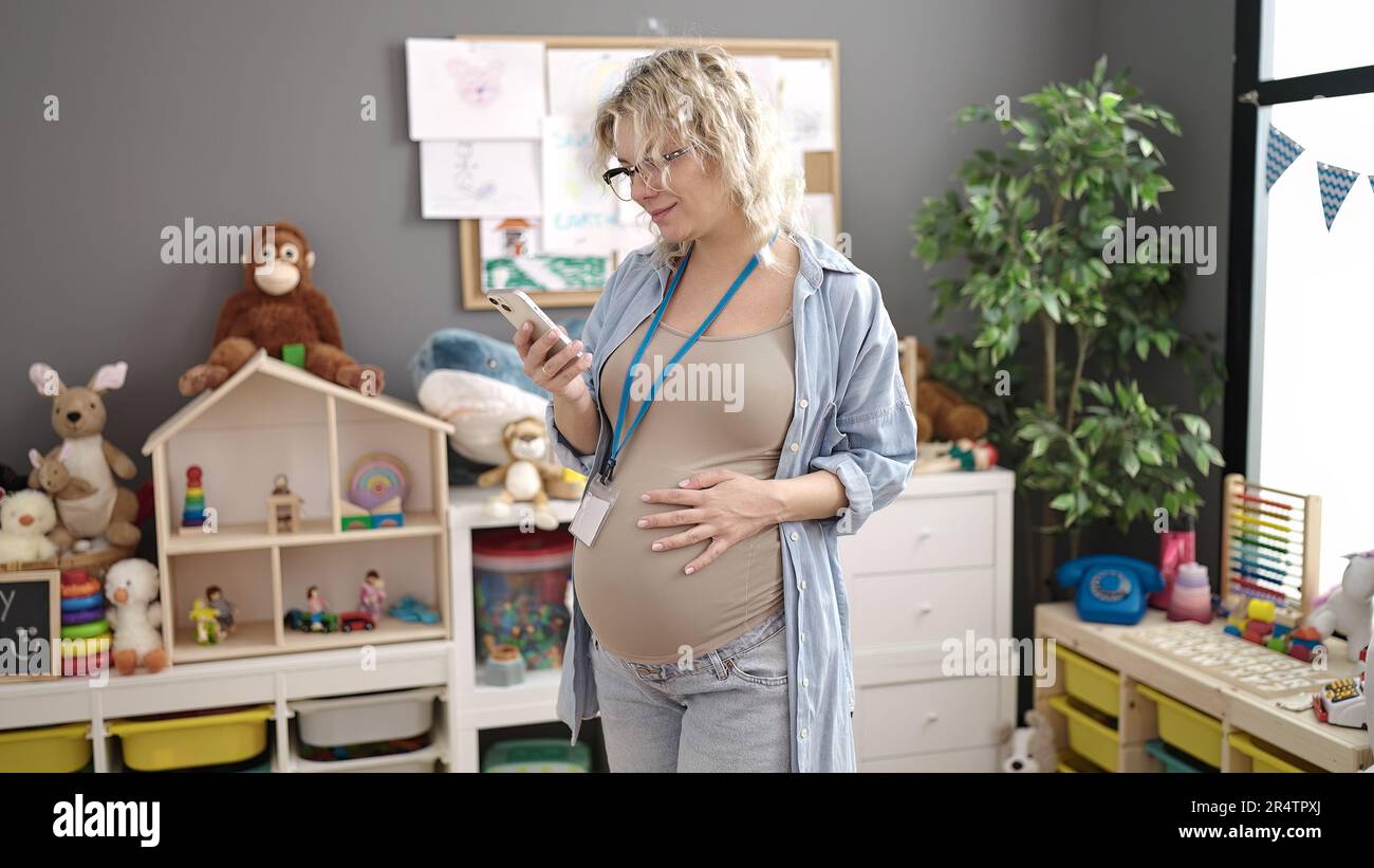 Junge schwangere Vorschullehrerin mit Smartphone, die im Kindergarten