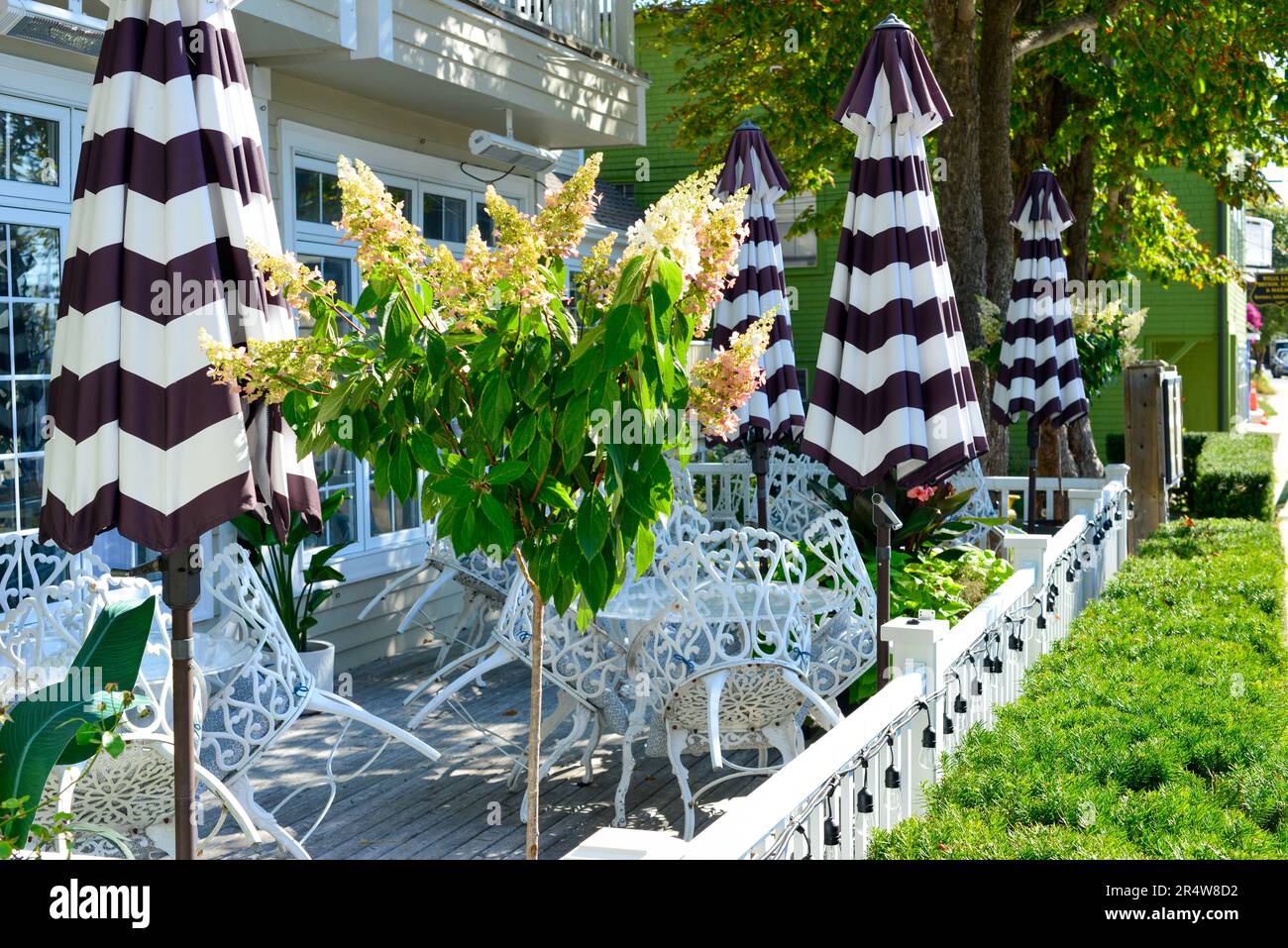 Mehrere leere weiße schmiedeeiserne Tische und Stühle auf der Terrasse des Hotelrestaurants. Es gibt schwarz-weiß gestreifte farbige Sonnenschirme und Hecken. Stockfoto