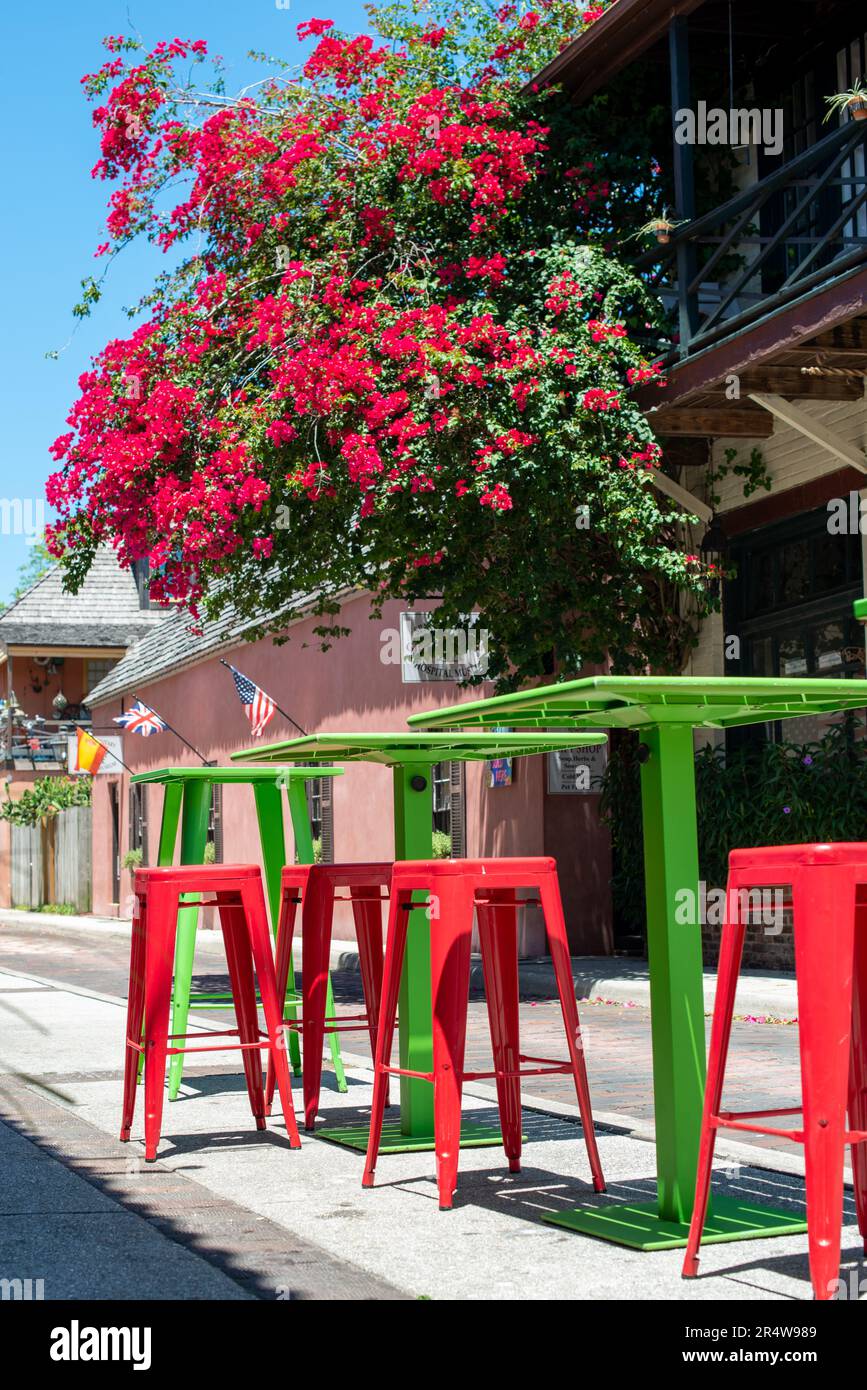 Auf einem Gehweg vor einem Café mit blühenden Bäumen und roten Blumen stehen dunkelrote und farbenfrohe grüne Metalltische und -Stühle in Pubgröße Stockfoto